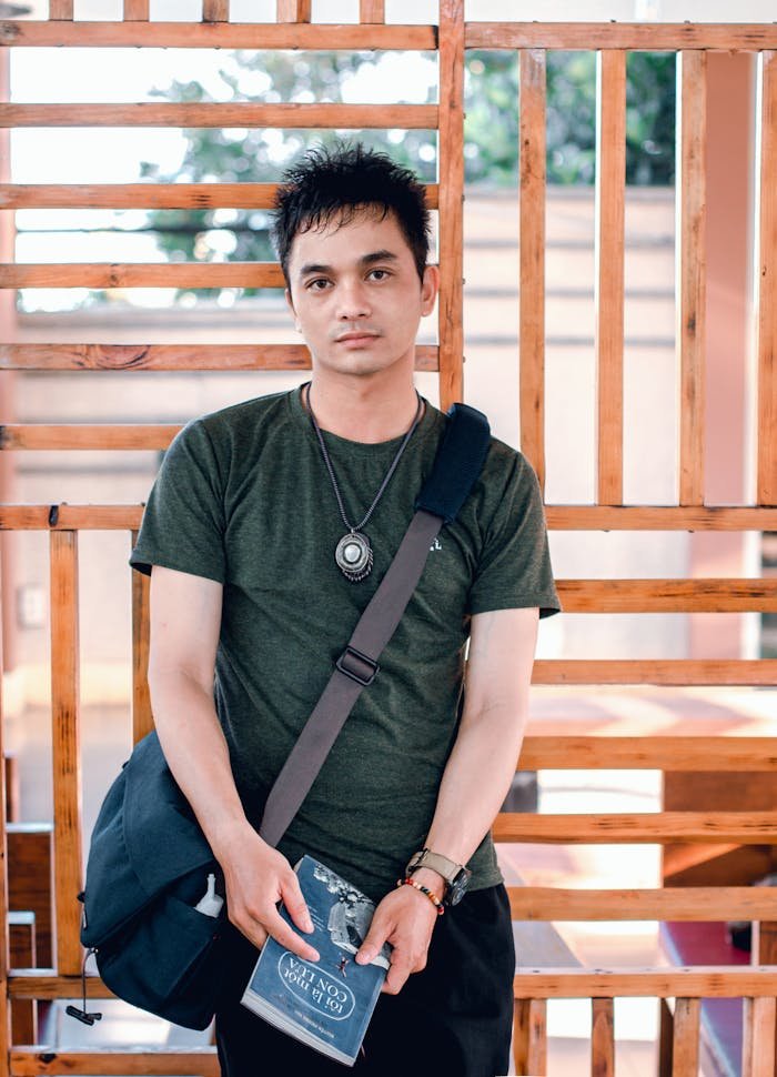 Young adult man holds a book and bag in a casual outdoor setting, appearing pensive.